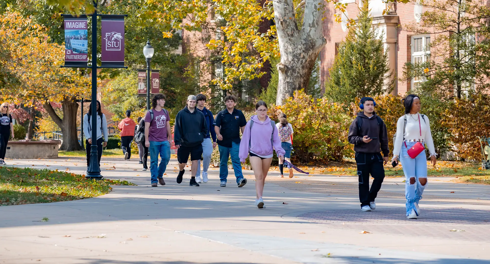 Students walking on a tree-lined campus pathway in autumn, with leaves scattered on the ground and a warm mix of green, yellow, and orange foliage. SIU banners are visible on lampposts, creating a welcoming and vibrant atmosphere.